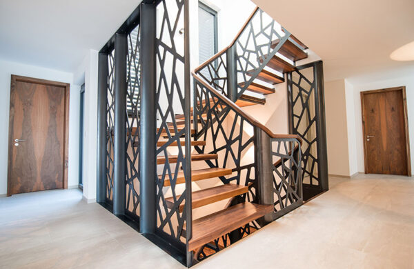 Treppe mit Holztritten und einem Metallgeländer mit Holzauflagen. Das Metall ist in schwarz mit eckigen Ausschnitten.